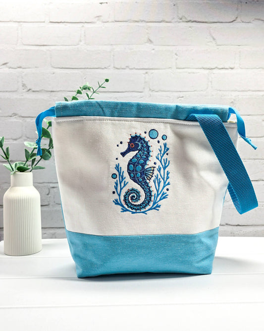 Projekttasche Tuva M aus hellem Canvas und hellblauem Stoff mit blau gesticktem Seepferdchen-Motiv im Korallenkranz, Kordelzug und blauem Tragegurt. Bietet Platz für 400g Wolle und schützt das Strickzeug vor Verheddern. Handgenäht in Bayern von Nadelmikado.