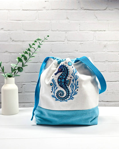 Projekttasche Tuva M aus hellem Canvas und hellblauem Stoff mit blau gesticktem Seepferdchen-Motiv im Korallenkranz, Kordelzug und blauem Tragegurt. Bietet Platz für 400g Wolle und schützt das Strickzeug vor Verheddern. Handgenäht in Bayern von Nadelmikado.