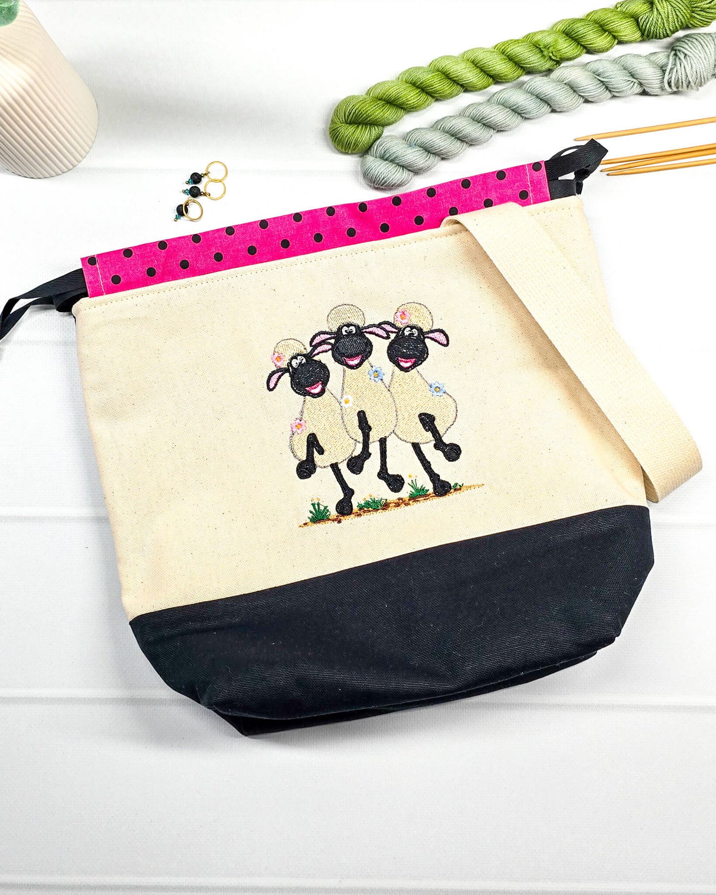Projekttasche Tuva M aus hellem Canvas und schwarzem Stoff mit drei gestickten, tanzenden Schafen, pink-schwarz gepunktetem Kordelzug-Bündchen und cremefarbenem Tragegurt. Bietet Platz für 400g Wolle und schützt das Strickzeug vor Verheddern. Handgenäht in Bayern von Nadelmikado.