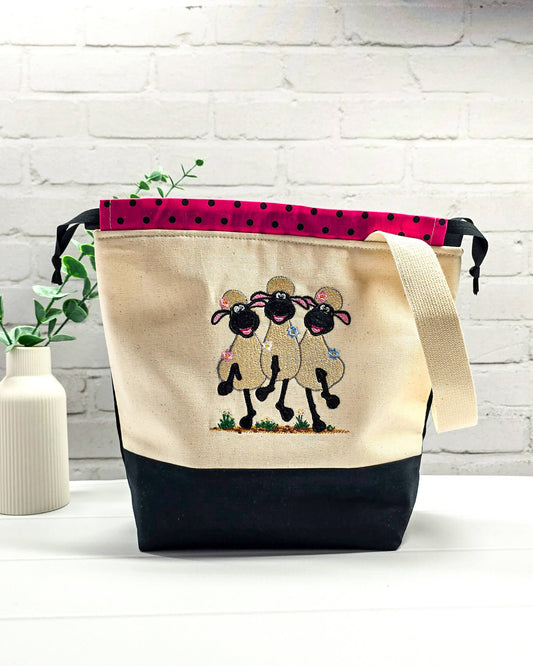 Projekttasche Tuva M aus hellem Canvas und schwarzem Stoff mit drei gestickten, tanzenden Schafen, pink-schwarz gepunktetem Kordelzug-Bündchen und cremefarbenem Tragegurt. Bietet Platz für 400g Wolle und schützt das Strickzeug vor Verheddern. Handgenäht in Bayern von Nadelmikado.
