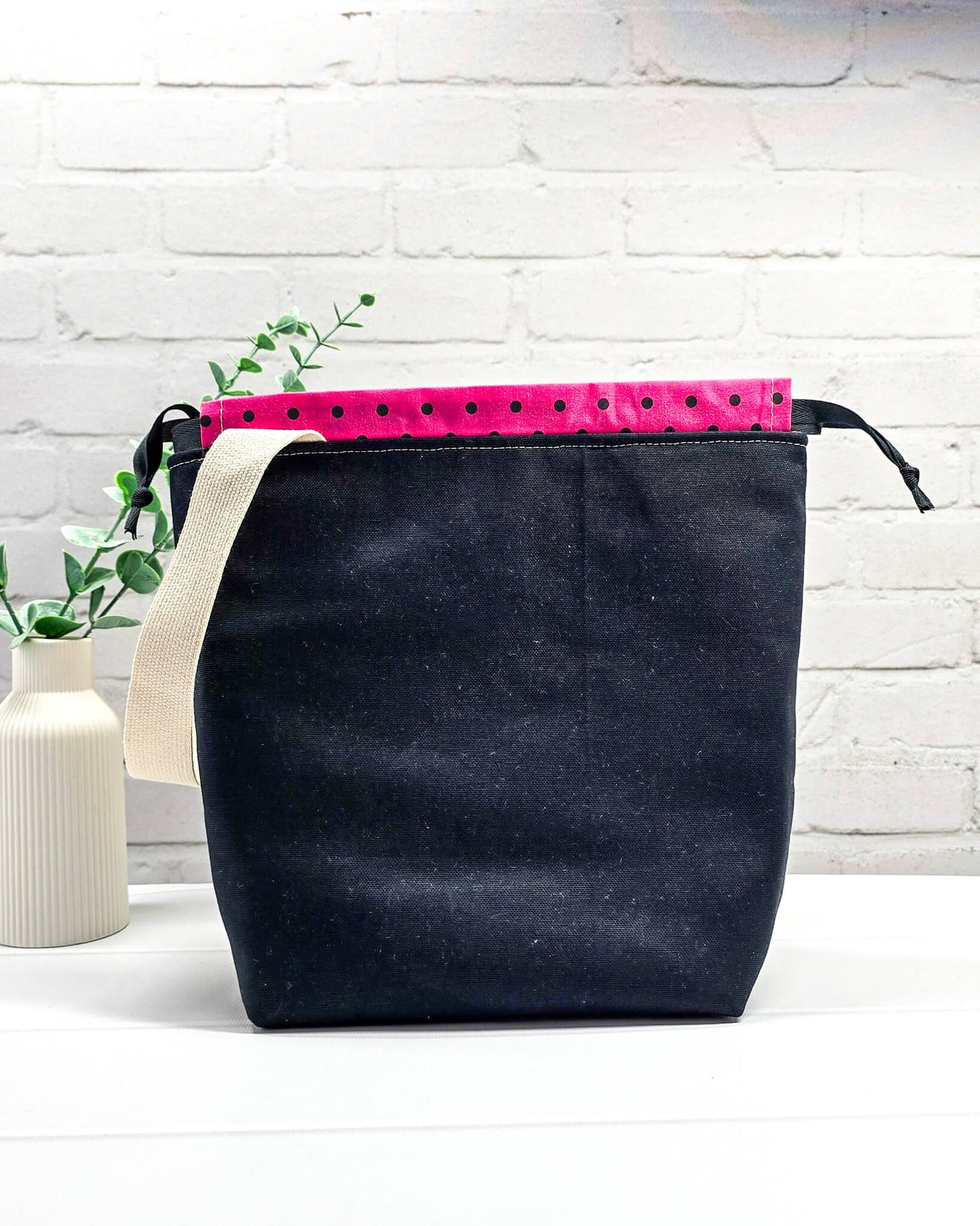 Projekttasche Tuva M aus hellem Canvas und schwarzem Stoff mit drei gestickten, tanzenden Schafen, pink-schwarz gepunktetem Kordelzug-Bündchen und cremefarbenem Tragegurt. Bietet Platz für 400g Wolle und schützt das Strickzeug vor Verheddern. Handgenäht in Bayern von Nadelmikado.