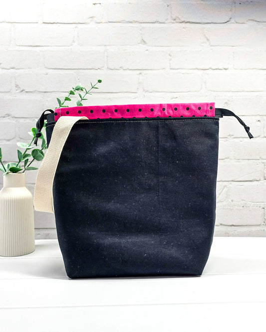 Projekttasche Tuva M aus hellem Canvas und schwarzem Stoff mit drei gestickten, tanzenden Schafen, pink-schwarz gepunktetem Kordelzug-Bündchen und cremefarbenem Tragegurt. Bietet Platz für 400g Wolle und schützt das Strickzeug vor Verheddern. Handgenäht in Bayern von Nadelmikado.