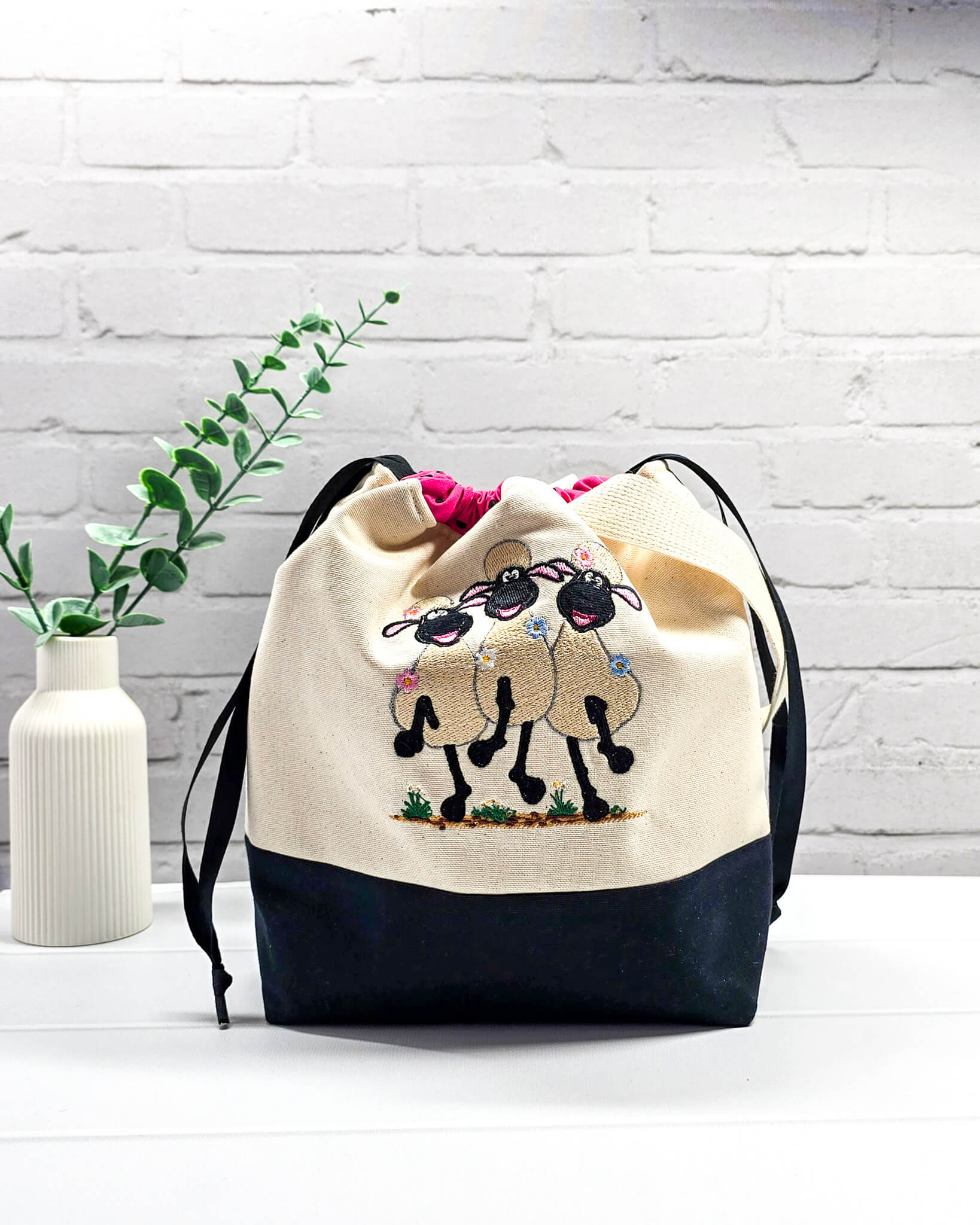 Projekttasche Tuva M aus hellem Canvas und schwarzem Stoff mit drei gestickten, tanzenden Schafen, pink-schwarz gepunktetem Kordelzug-Bündchen und cremefarbenem Tragegurt. Bietet Platz für 400g Wolle und schützt das Strickzeug vor Verheddern. Handgenäht in Bayern von Nadelmikado.