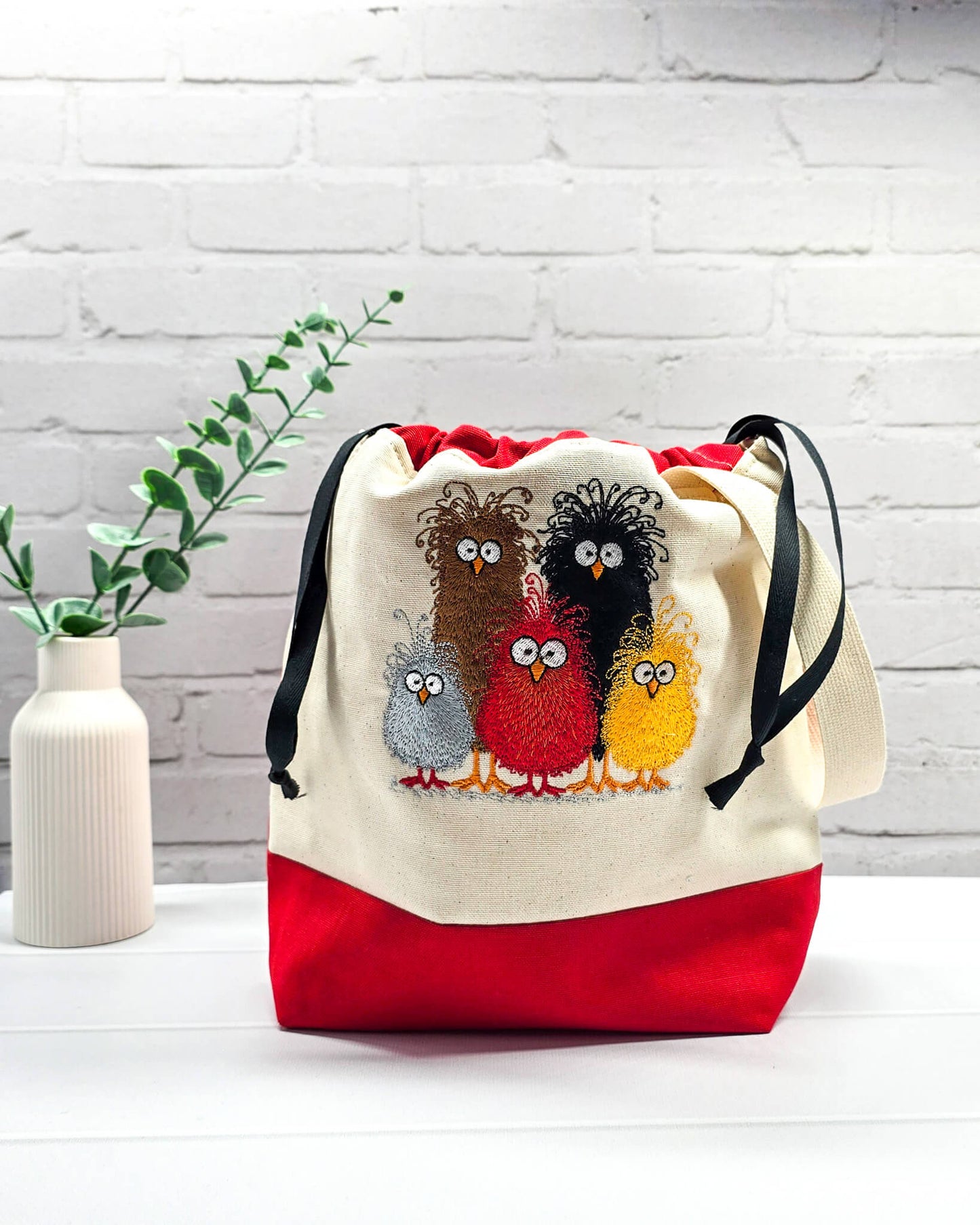 Projekttasche Tuva M aus hellem Canvas und rotem Stoff mit fünf bunt gestickten, strubbeligen Vögeln, schwarzem Kordelzug und cremefarbenem Tragegurt. Bietet Platz für 400g Wolle und schützt das Strickzeug vor Verheddern. Handgenäht in Bayern von Nadelmikado.