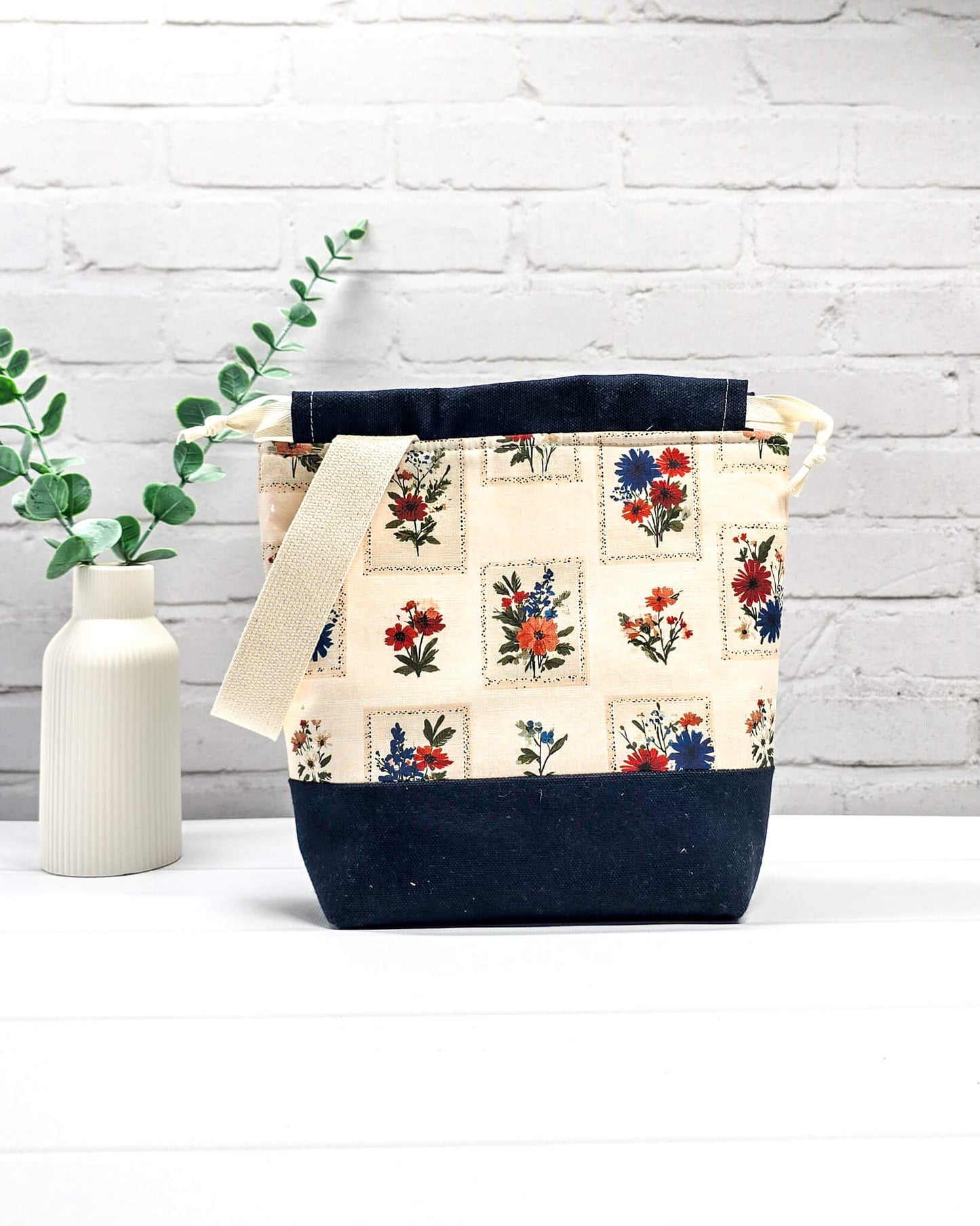Handgenähter Projektbeutel mit floralem Motiv aus eingerahmten Wiesenblumen auf hellem Grund, dunklem Boden, cremefarbenem Kordelzug und Tragegurt. Für 200g Wolle. Handgenäht in Bayern von Nadelmikado.