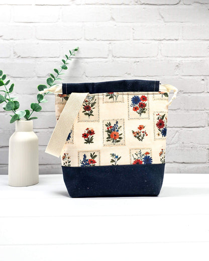 Handgenähter Projektbeutel mit floralem Motiv aus eingerahmten Wiesenblumen auf hellem Grund, dunklem Boden, cremefarbenem Kordelzug und Tragegurt. Für 200g Wolle. Handgenäht in Bayern von Nadelmikado.
