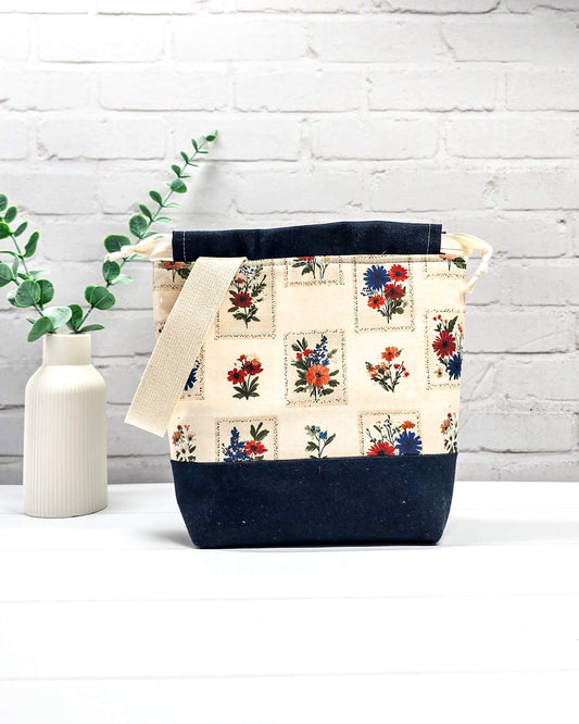 Handgenähter Projektbeutel mit floralem Motiv aus eingerahmten Wiesenblumen auf hellem Grund, dunklem Boden, cremefarbenem Kordelzug und Tragegurt. Für 200g Wolle. Handgenäht in Bayern von Nadelmikado.
