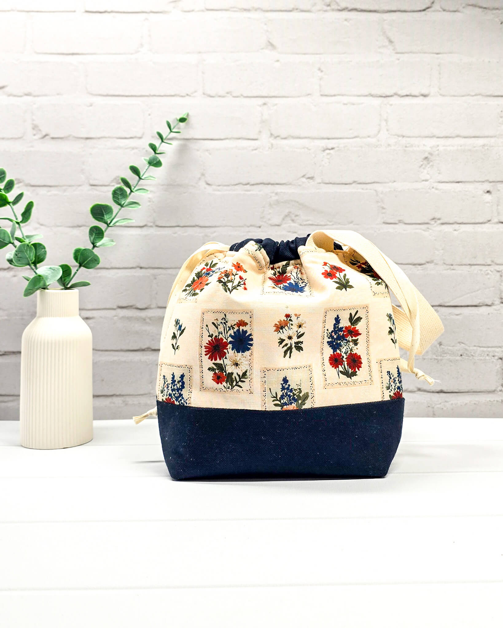 Handgenähter Projektbeutel mit floralem Motiv aus eingerahmten Wiesenblumen auf hellem Grund, dunklem Boden, cremefarbenem Kordelzug und Tragegurt. Für 200g Wolle. Handgenäht in Bayern von Nadelmikado.