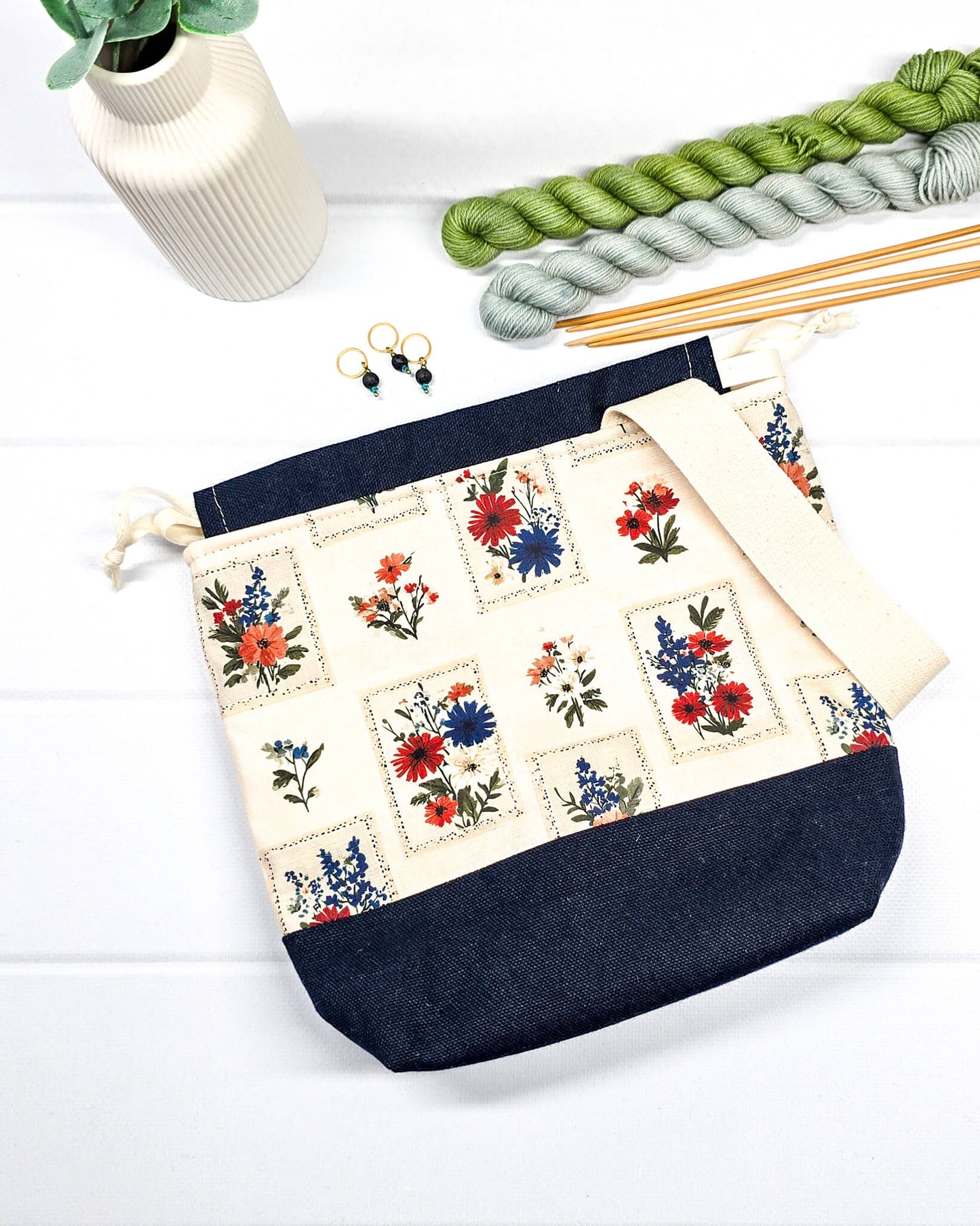 Handgenähter Projektbeutel mit floralem Motiv aus eingerahmten Wiesenblumen auf hellem Grund, dunklem Boden, cremefarbenem Kordelzug und Tragegurt. Für 200g Wolle. Handgenäht in Bayern von Nadelmikado.