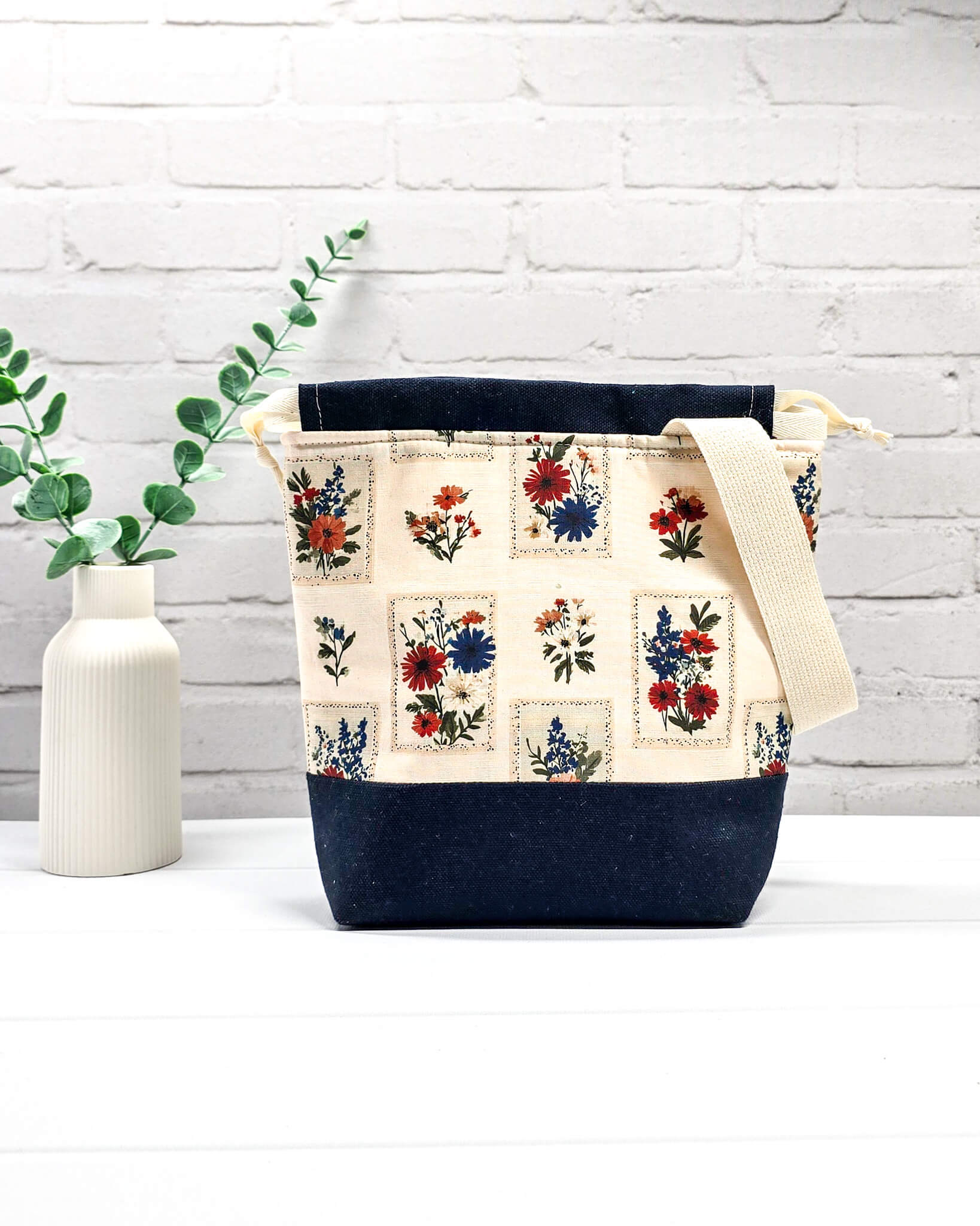 Handgenähter Projektbeutel mit floralem Motiv aus eingerahmten Wiesenblumen auf hellem Grund, dunklem Boden, cremefarbenem Kordelzug und Tragegurt. Für 200g Wolle. Handgenäht in Bayern von Nadelmikado.