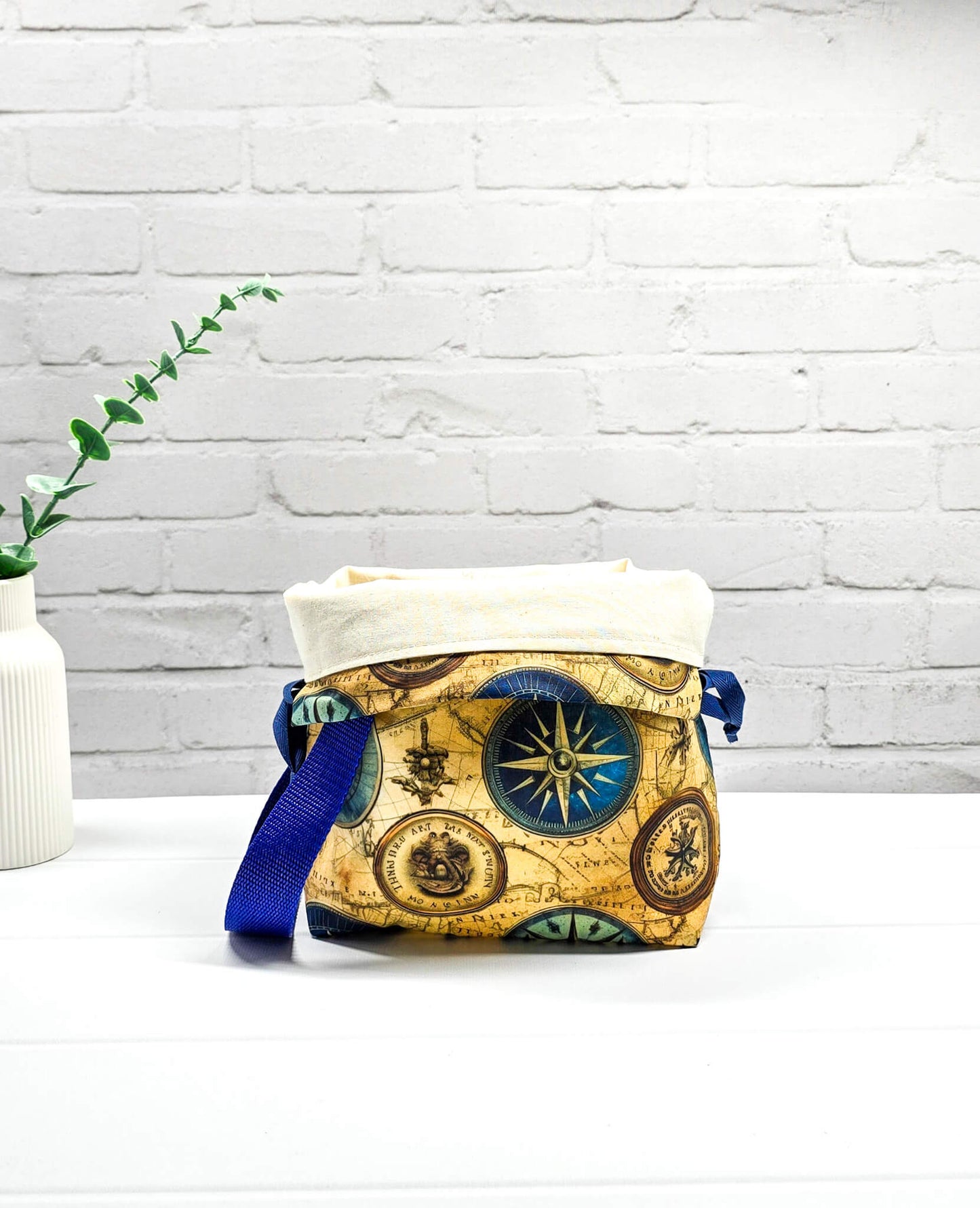 Handgenähte Projekttasche Tuva S von Nadelmikado mit Kompass-Motiv und Vintage-Weltkarte, seitlicher blauer Tragegurt, stehend auf weißem Grund.