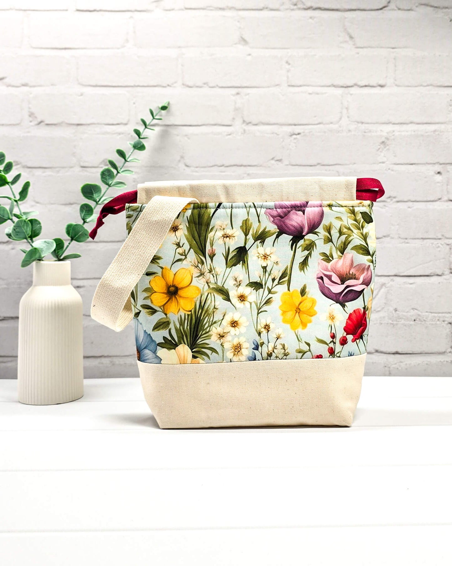Handgenähter Projektbeutel mit buntem Blumenmotiv auf hellblauem Grund, beigem Boden, roter Kordel und hellem Tragegurt. Für 200g Wolle. Handgenäht in Bayern von Nadelmikado.