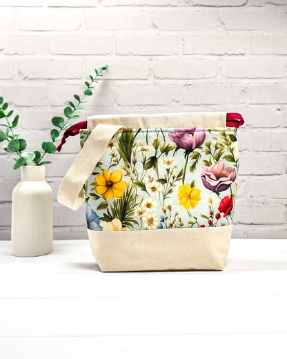 Handgenähter Projektbeutel mit buntem Blumenmotiv auf hellblauem Grund, beigem Boden, roter Kordel und hellem Tragegurt. Für 200g Wolle. Handgenäht in Bayern von Nadelmikado.
