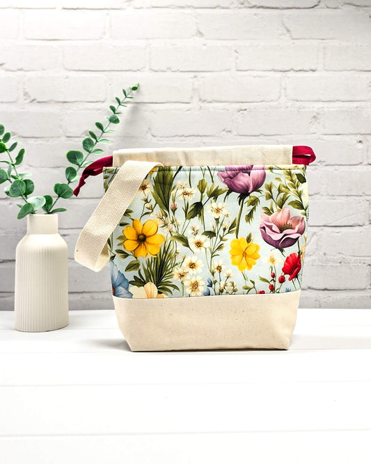 Handgenähter Projektbeutel mit buntem Blumenmotiv auf hellblauem Grund, beigem Boden, roter Kordel und hellem Tragegurt. Für 200g Wolle. Handgenäht in Bayern von Nadelmikado.