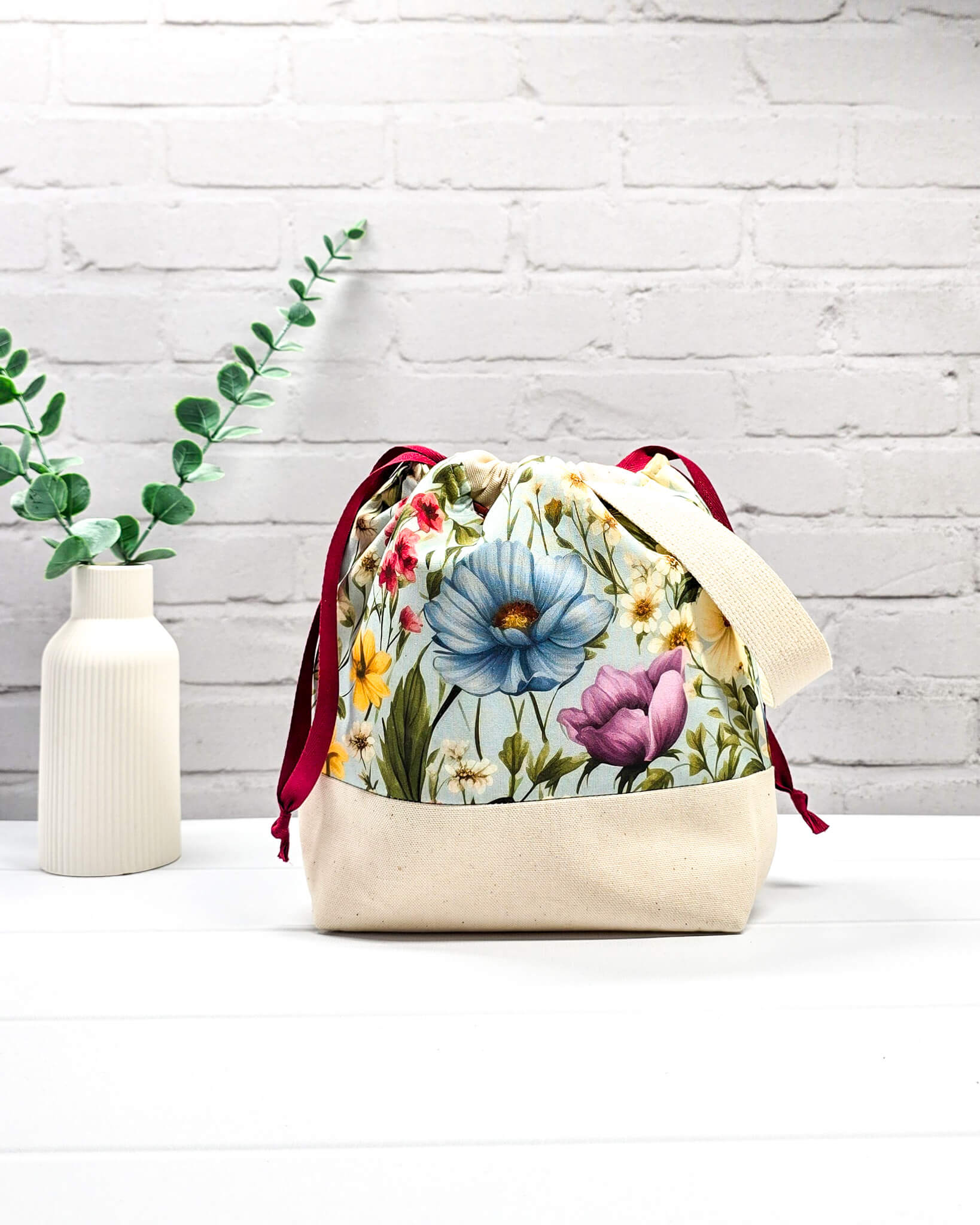 Handgenähter Projektbeutel mit buntem Blumenmotiv auf hellblauem Grund, beigem Boden, roter Kordel und hellem Tragegurt. Für 200g Wolle. Handgenäht in Bayern von Nadelmikado.