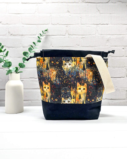 Kleine Handarbeitstasche mit Katzen-Motiv auf dunklem Grund, schwarzem Boden, schwarzem Kordelzug und hellem Tragegurt. Für 200g Wolle. Handgenäht in Bayern von Nadelmikado.