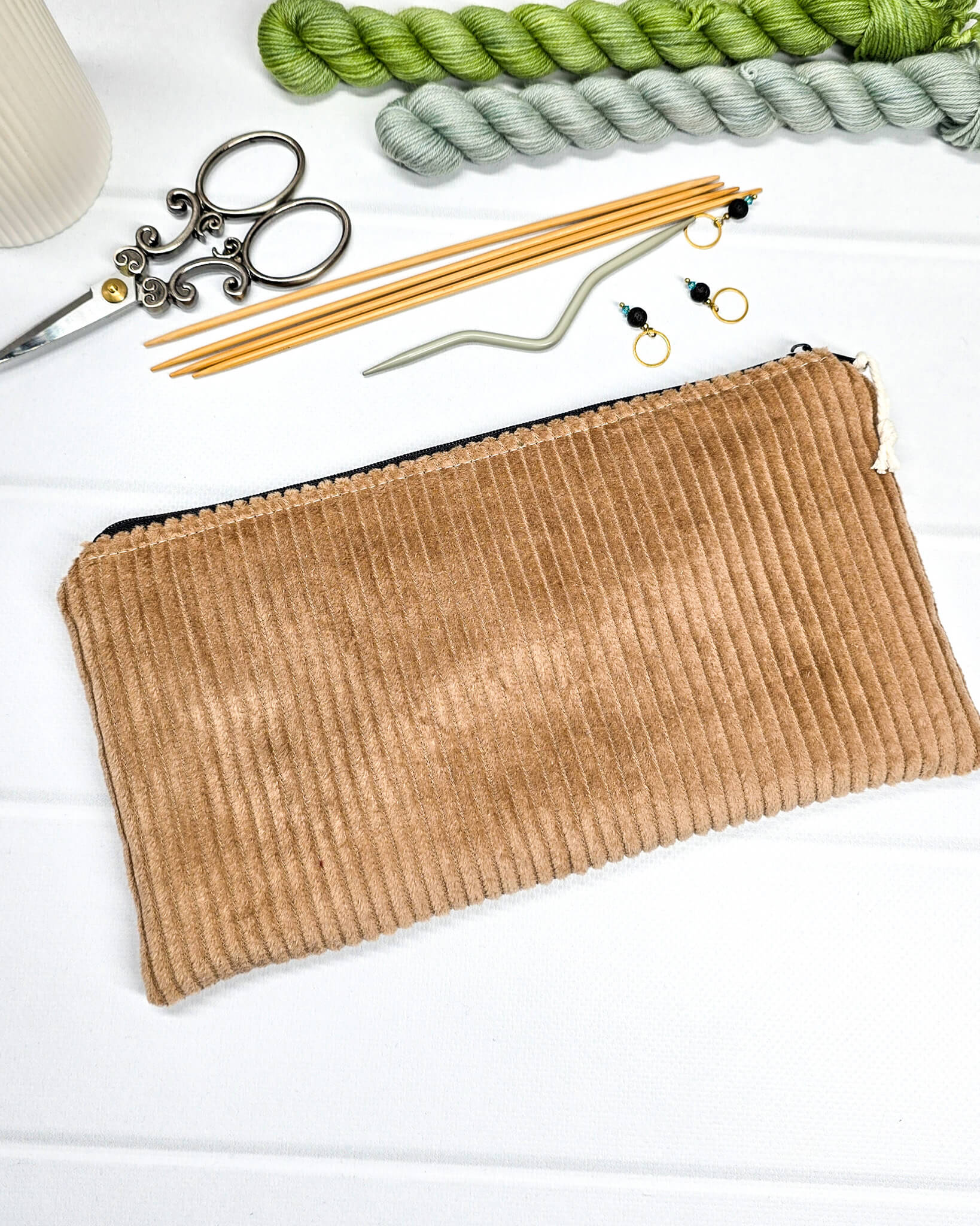 Kleine camelbraune Reißverschlusstasche aus Cord-Stoff für Strickzubehör, liegend auf weißem Untergrund mit einer Schere, Garnsträngen und Holznadeln. Handgenäht in Bayern von Nadelmikado.