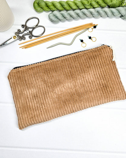 Kleine camelbraune Reißverschlusstasche aus Cord-Stoff für Strickzubehör, liegend auf weißem Untergrund mit einer Schere, Garnsträngen und Holznadeln. Handgenäht in Bayern von Nadelmikado.