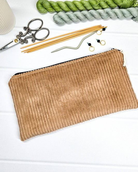 Kleine camelbraune Reißverschlusstasche aus Cord-Stoff für Strickzubehör, liegend auf weißem Untergrund mit einer Schere, Garnsträngen und Holznadeln. Handgenäht in Bayern von Nadelmikado.