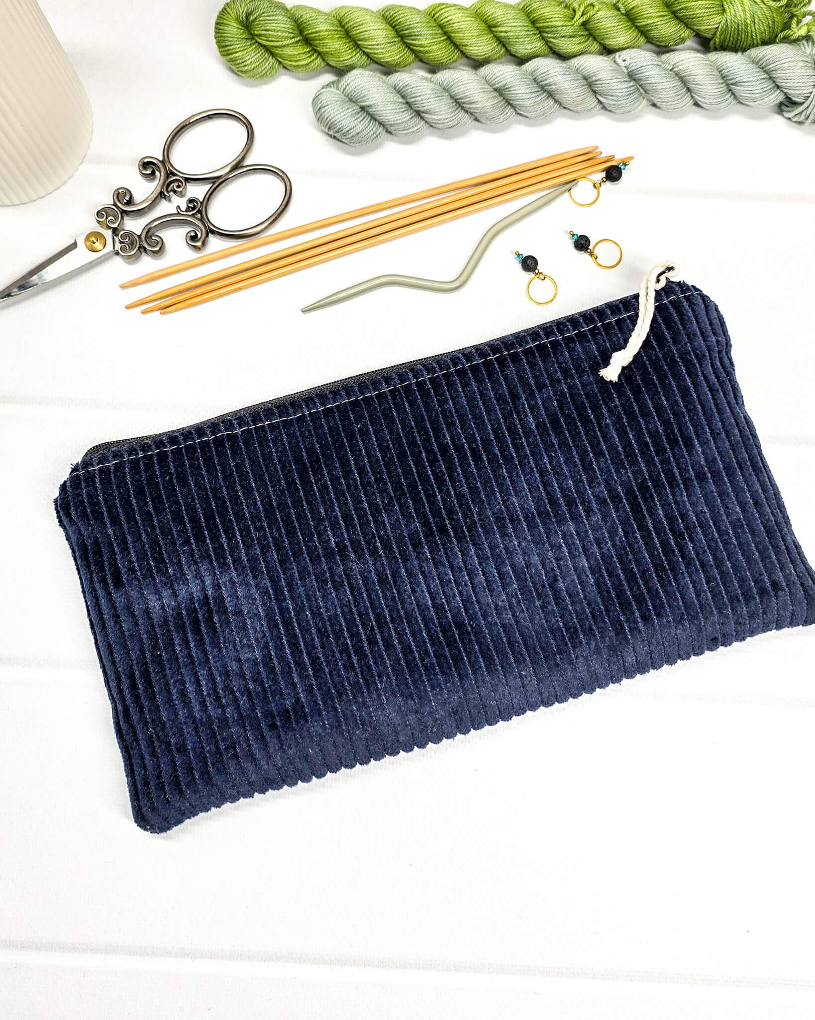 Kleine Reißverschlusstasche aus dunkelblauem Cord-Stoff für Strickzubehör, flach liegend auf weißem Untergrund mit Schere, Garn und Nadeln. Handgenäht in Bayern von Nadelmikado.