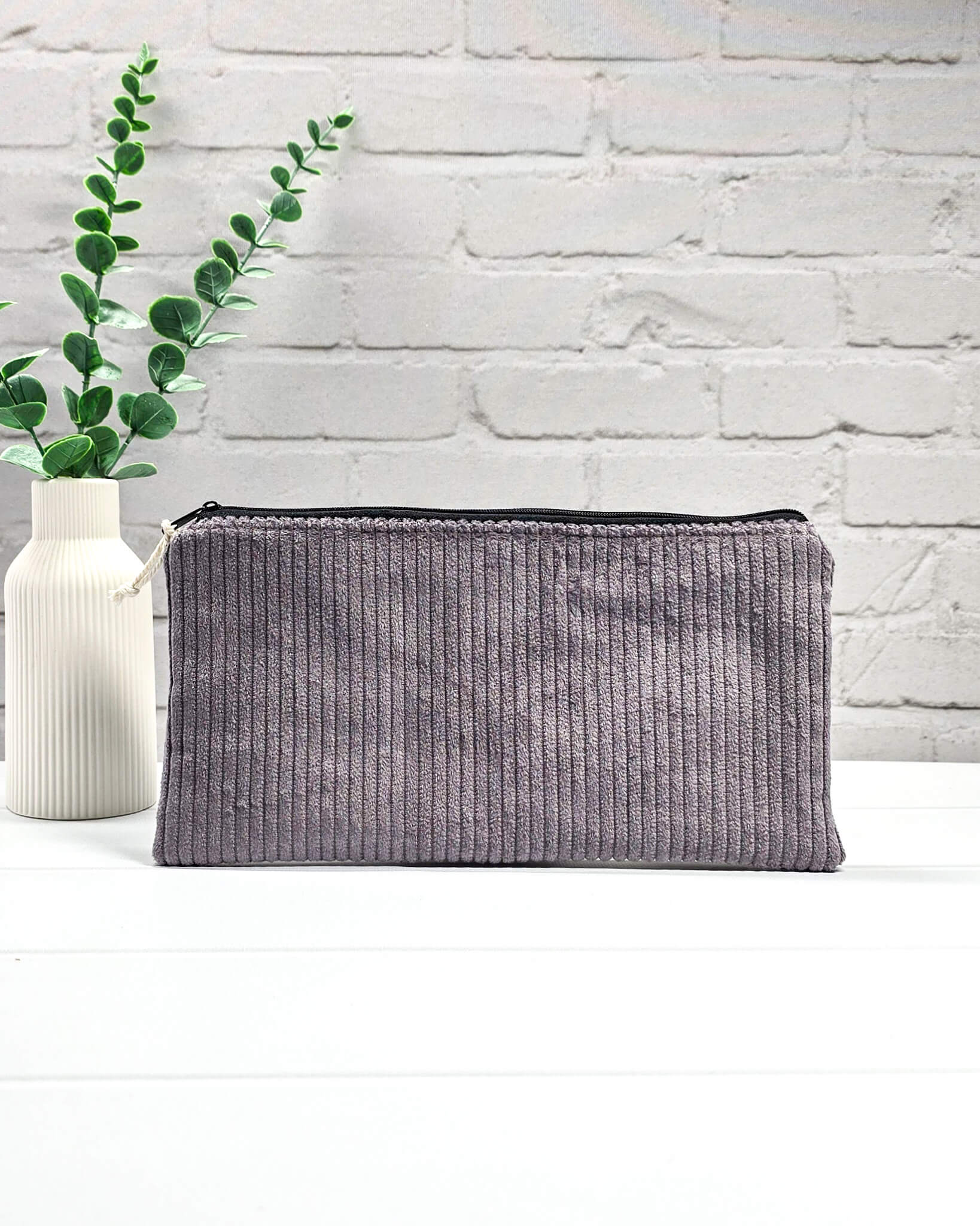 Kleine graulila Reißverschlusstasche aus Cord-Stoff für Strickzubehör, liegend auf weißem Untergrund mit einer Schere, Garnsträngen und Holznadeln. Handgenäht in Bayern von Nadelmikado.