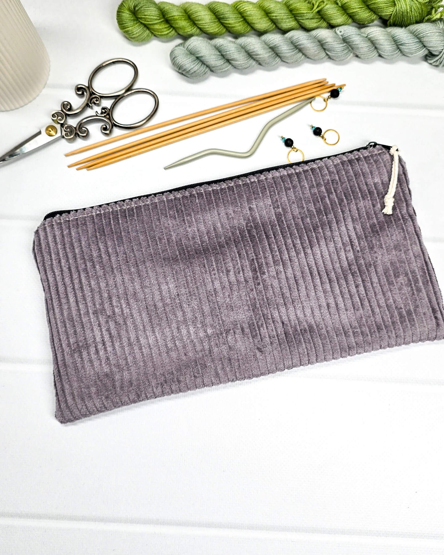 Kleine graulila Reißverschlusstasche aus Cord-Stoff für Strickzubehör, liegend auf weißem Untergrund mit einer Schere, Garnsträngen und Holznadeln. Handgenäht in Bayern von Nadelmikado.