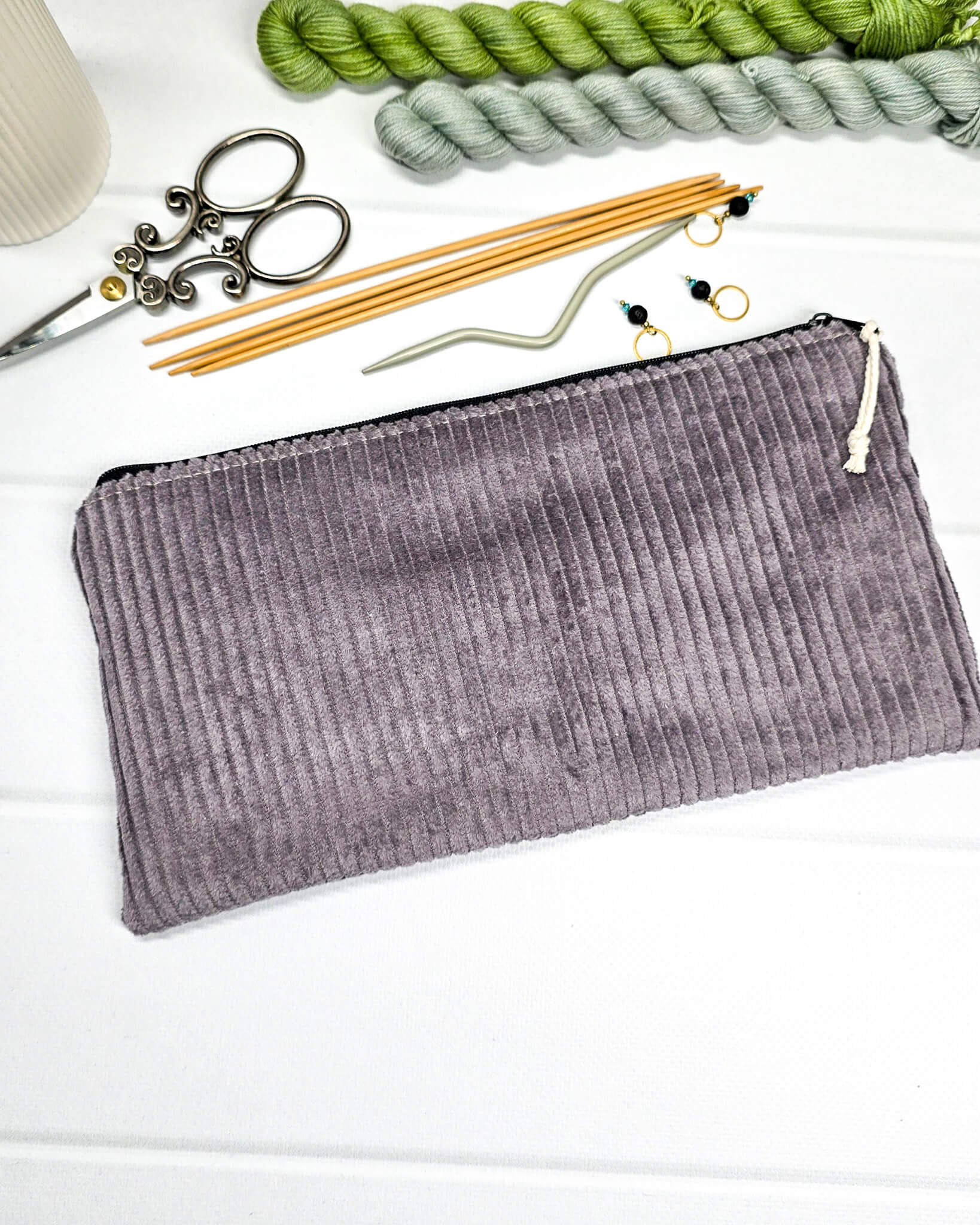 Kleine graulila Reißverschlusstasche aus Cord-Stoff für Strickzubehör, liegend auf weißem Untergrund mit einer Schere, Garnsträngen und Holznadeln. Handgenäht in Bayern von Nadelmikado.