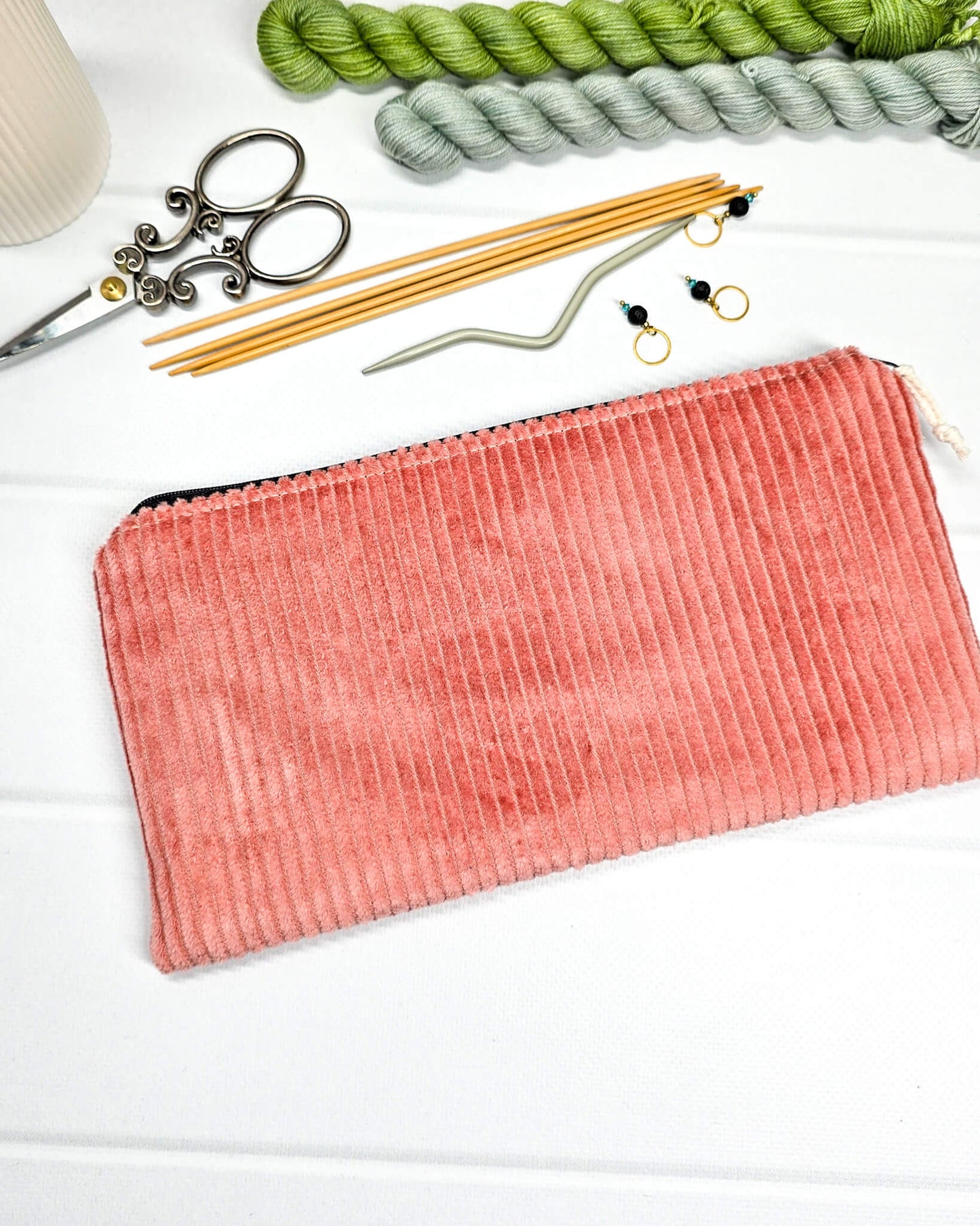 Kleine lachsfarbene Reißverschlusstasche aus Cord-Stoff für Strickzubehör, liegend auf weißem Untergrund mit einer Schere, Garnsträngen und Holznadeln. Handgenäht in Bayern von Nadelmikado.