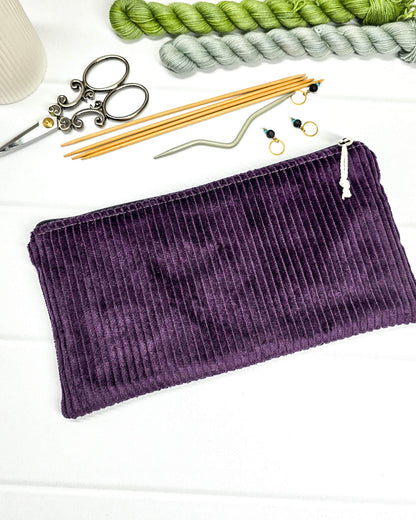 Kleine lila Reißverschlusstasche aus Cord-Stoff für Strickzubehör, flach liegend auf weißem Untergrund mit einer Schere, Garnsträngen und Holznadeln. Handgenäht in Bayern von Nadelmikado.