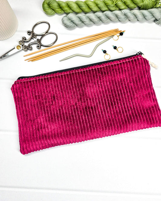 Kleine magentafarbene Reißverschlusstasche aus Cord-Stoff für Strickzubehör, liegend auf weißem Untergrund mit einer Schere, Garnsträngen und Holznadeln. Handgenäht in Bayern von Nadelmikado.
