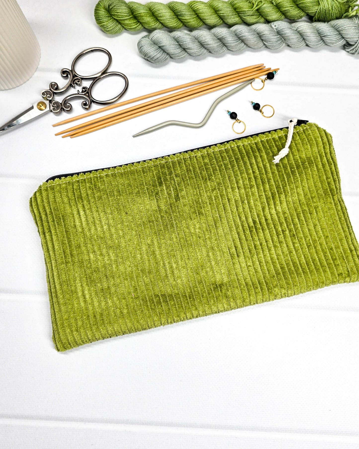 Kleine maigrüne Reißverschlusstasche aus Cord-Stoff für Strickzubehör, liegend auf weißem Untergrund mit einer Schere, Garnsträngen und Holznadeln. Handgenäht in Bayern von Nadelmikado.
