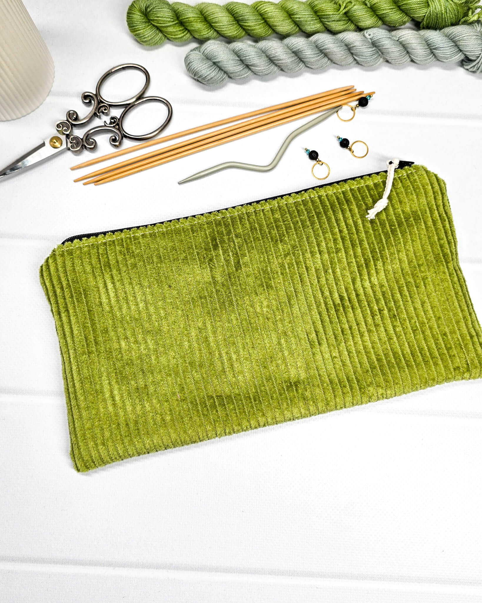 Kleine maigrüne Reißverschlusstasche aus Cord-Stoff für Strickzubehör, liegend auf weißem Untergrund mit einer Schere, Garnsträngen und Holznadeln. Handgenäht in Bayern von Nadelmikado.