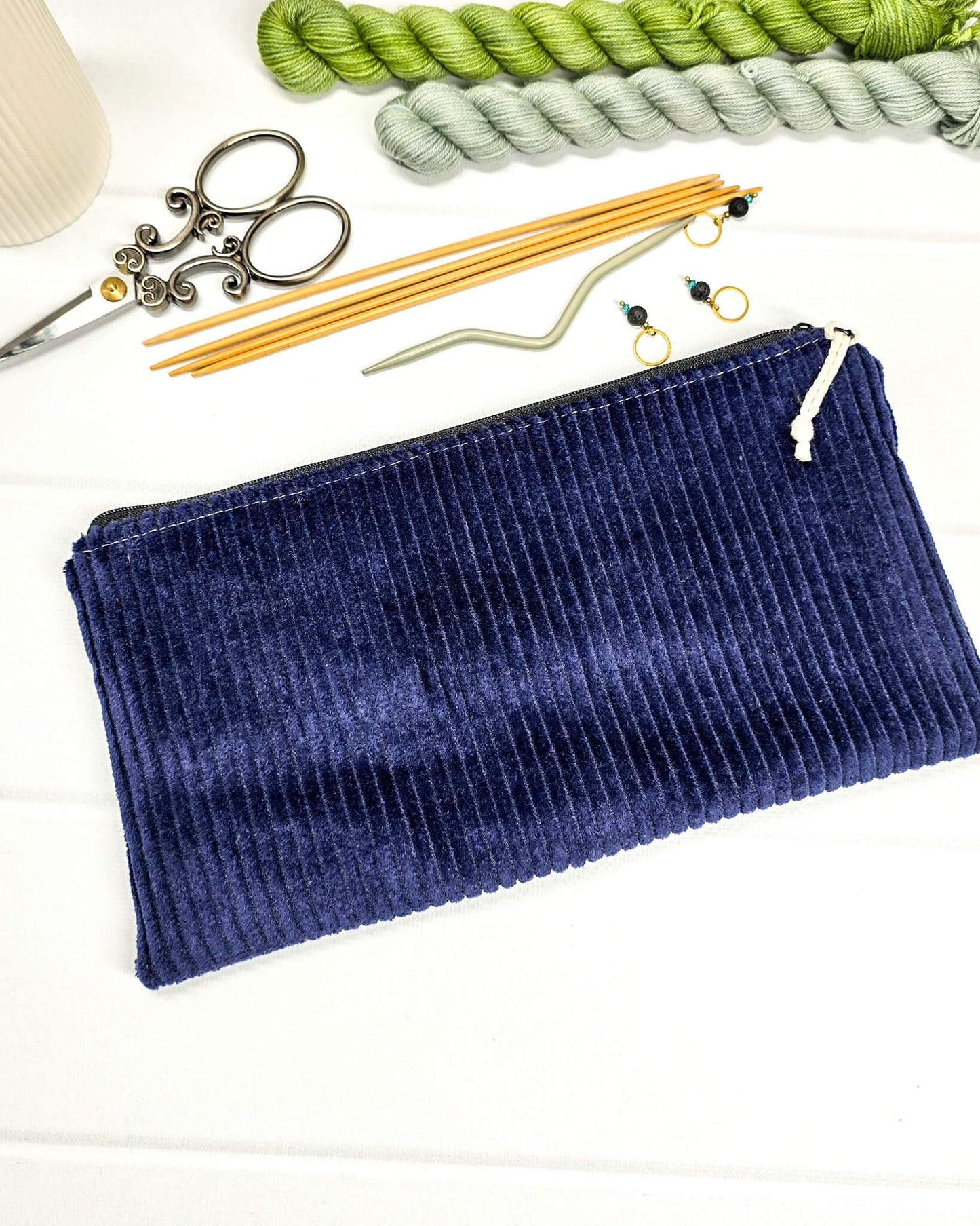 Kleine navyblaue Reißverschlusstasche aus Cord-Stoff für Strickzubehör, liegend auf weißem Untergrund mit einer Schere, Garnsträngen und Holznadeln. Handgenäht in Bayern von Nadelmikado.