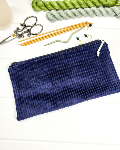 Kleine navyblaue Reißverschlusstasche aus Cord-Stoff für Strickzubehör, liegend auf weißem Untergrund mit einer Schere, Garnsträngen und Holznadeln. Handgenäht in Bayern von Nadelmikado.