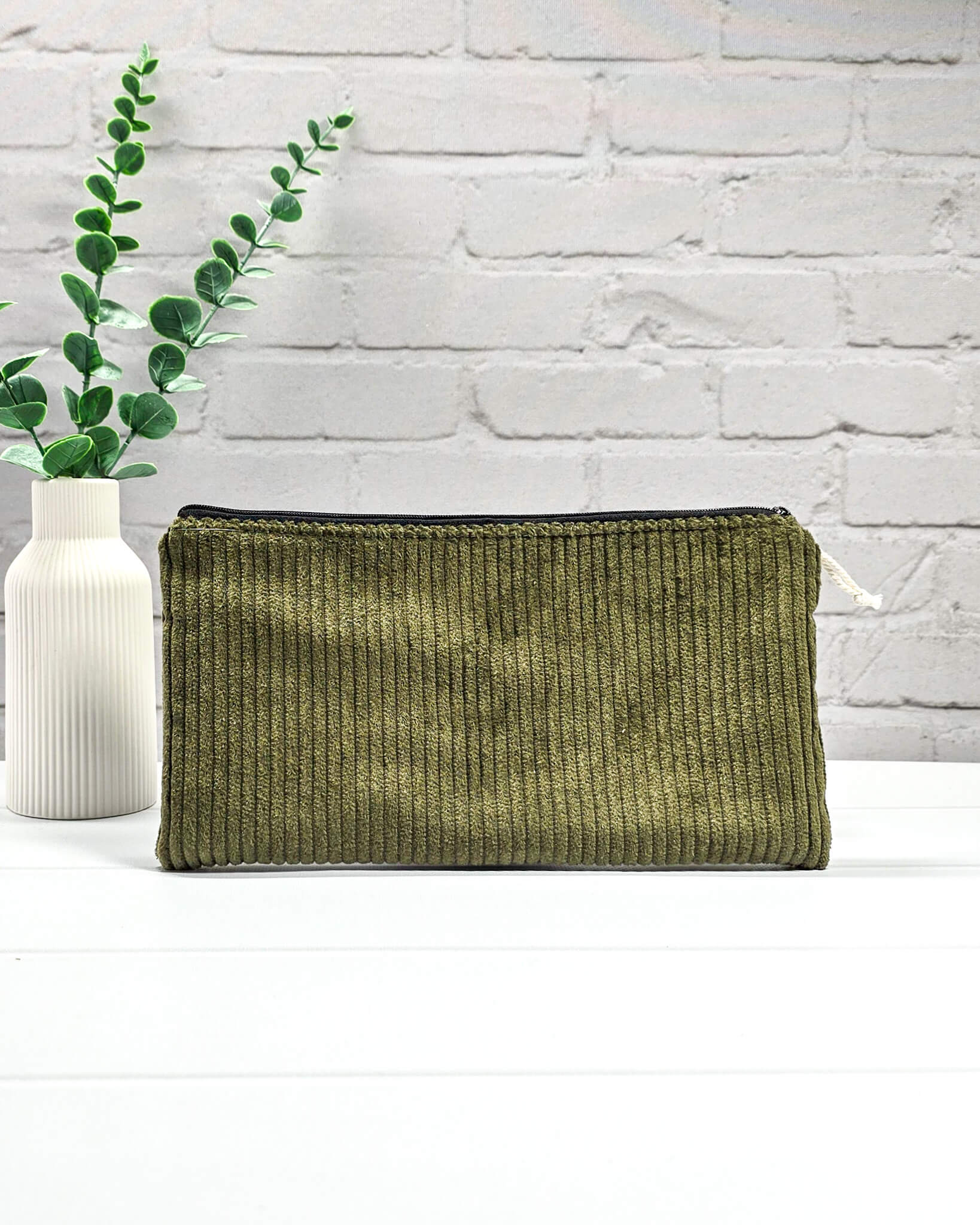 Kleine olivgrüne Reißverschlusstasche aus Cord-Stoff für Strickzubehör, liegend auf weißem Untergrund mit einer Schere, Garnsträngen und Holznadeln. Handgenäht in Bayern von Nadelmikado.