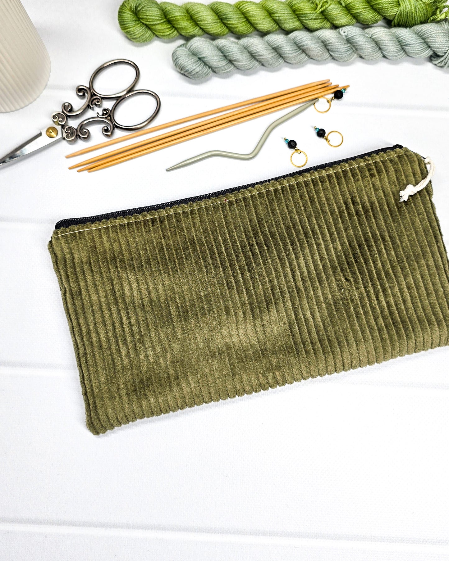 Kleine olivgrüne Reißverschlusstasche aus Cord-Stoff für Strickzubehör, liegend auf weißem Untergrund mit einer Schere, Garnsträngen und Holznadeln. Handgenäht in Bayern von Nadelmikado.