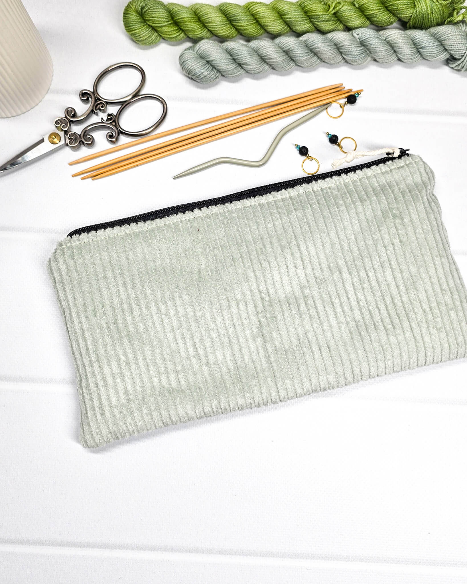Kleine Reißverschlusstasche aus hellgrünem Cord-Stoff für Strickzubehör, flach liegend auf weißem Untergrund mit Schere, Garn und Nadeln. Handgenäht in Bayern von Nadelmikado.