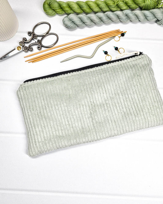 Kleine Reißverschlusstasche aus hellgrünem Cord-Stoff für Strickzubehör, flach liegend auf weißem Untergrund mit Schere, Garn und Nadeln. Handgenäht in Bayern von Nadelmikado.