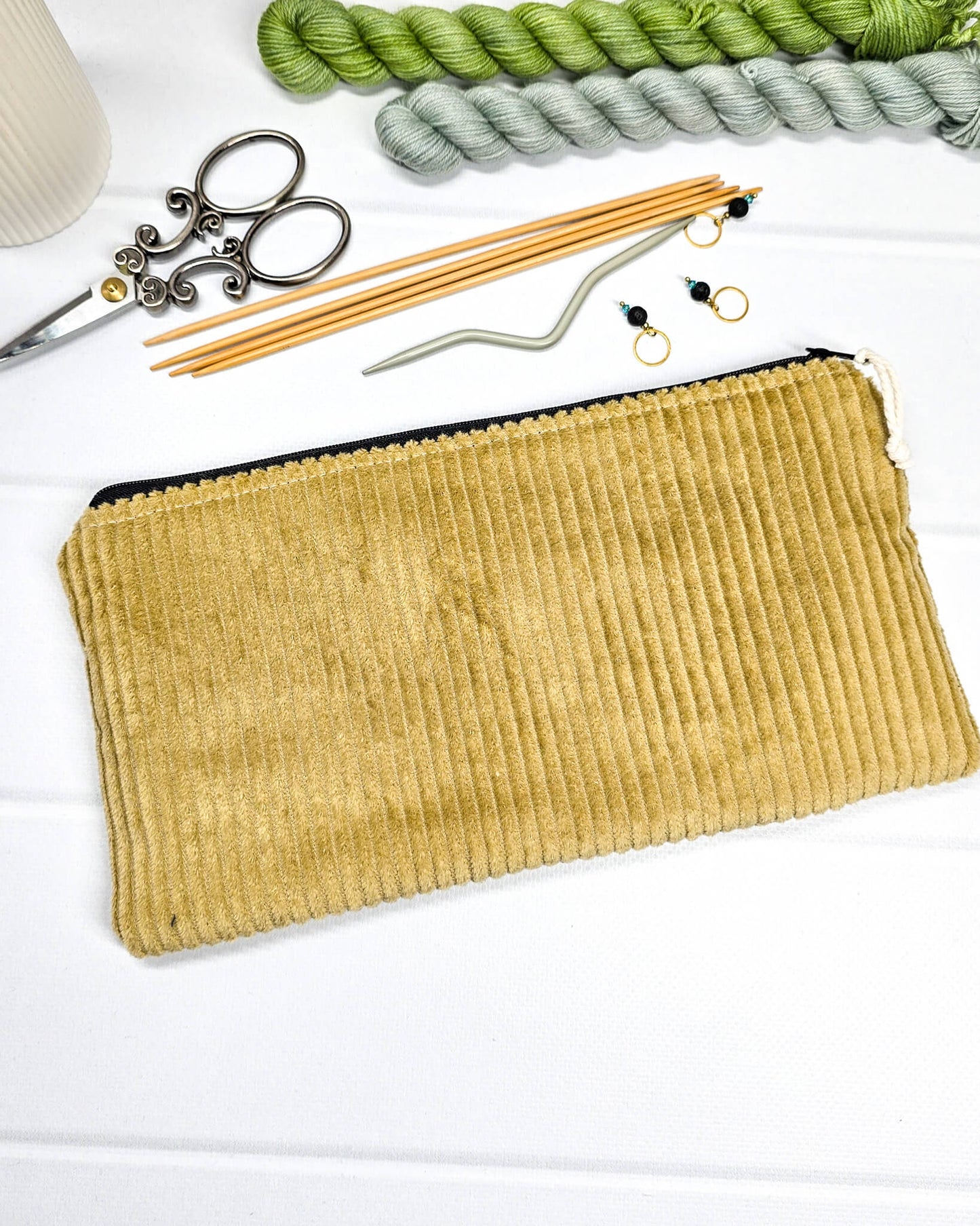 Kleine Reißverschlusstasche aus senfgelbem Cord-Stoff für Strickzubehör, flach liegend auf weißem Untergrund mit Schere, Garn und Nadeln. Handgenäht in Bayern von Nadelmikado.