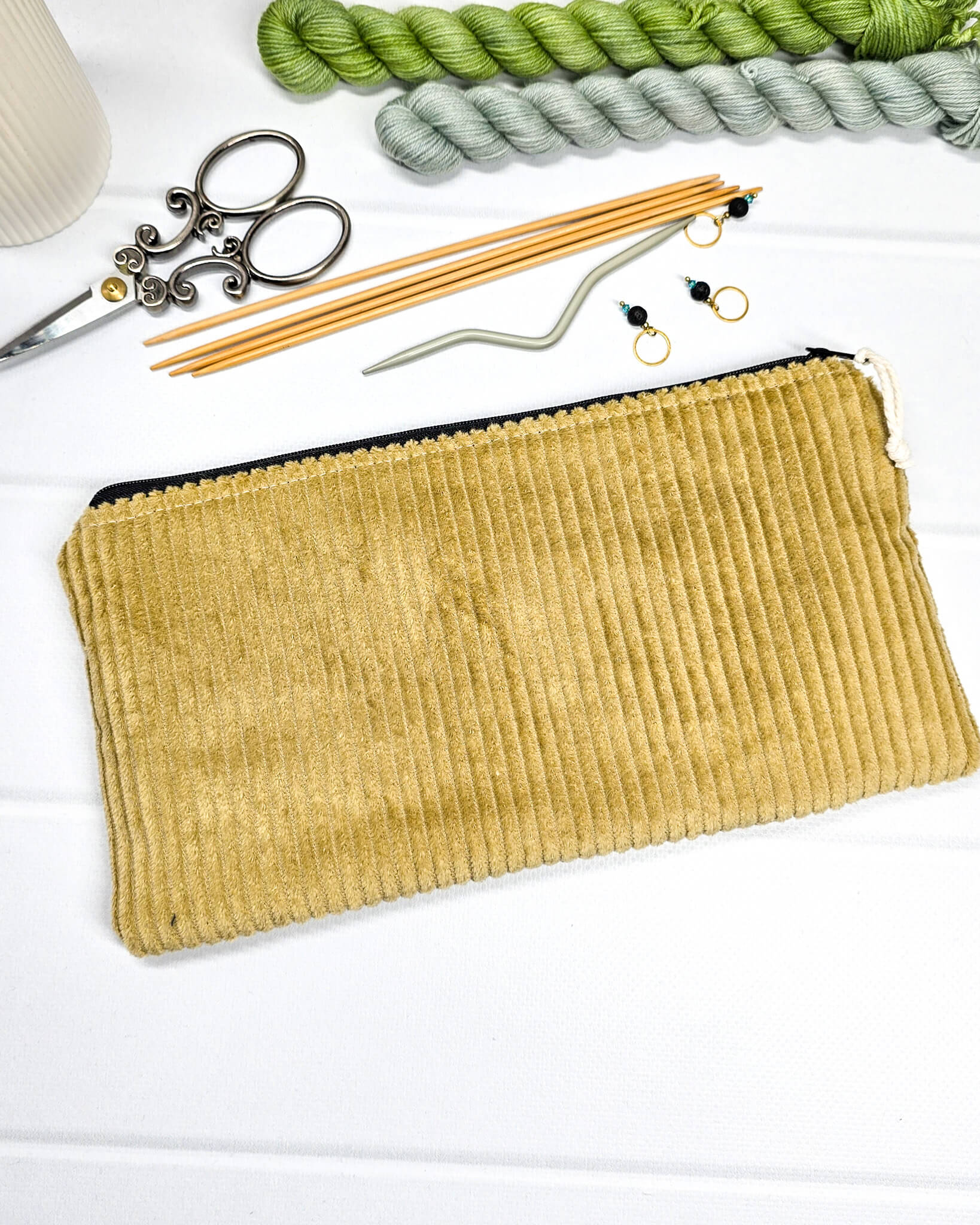 Kleine Reißverschlusstasche aus senfgelbem Cord-Stoff für Strickzubehör, flach liegend auf weißem Untergrund mit Schere, Garn und Nadeln. Handgenäht in Bayern von Nadelmikado.