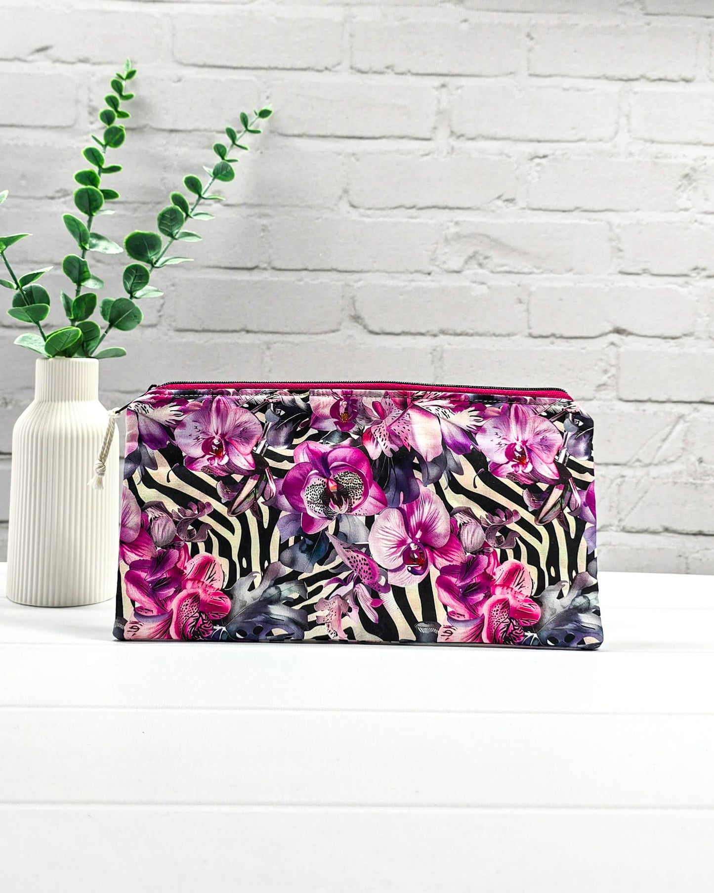 Längliche Tasche mit pinkfarbenem Reißverschluss, hellem Kordel-Zipper und auffälligem Muster aus lila Orchideenblüten über einem schwarz-weißen Zebramuster. Handgenäht in Bayern von Nadelmikado.