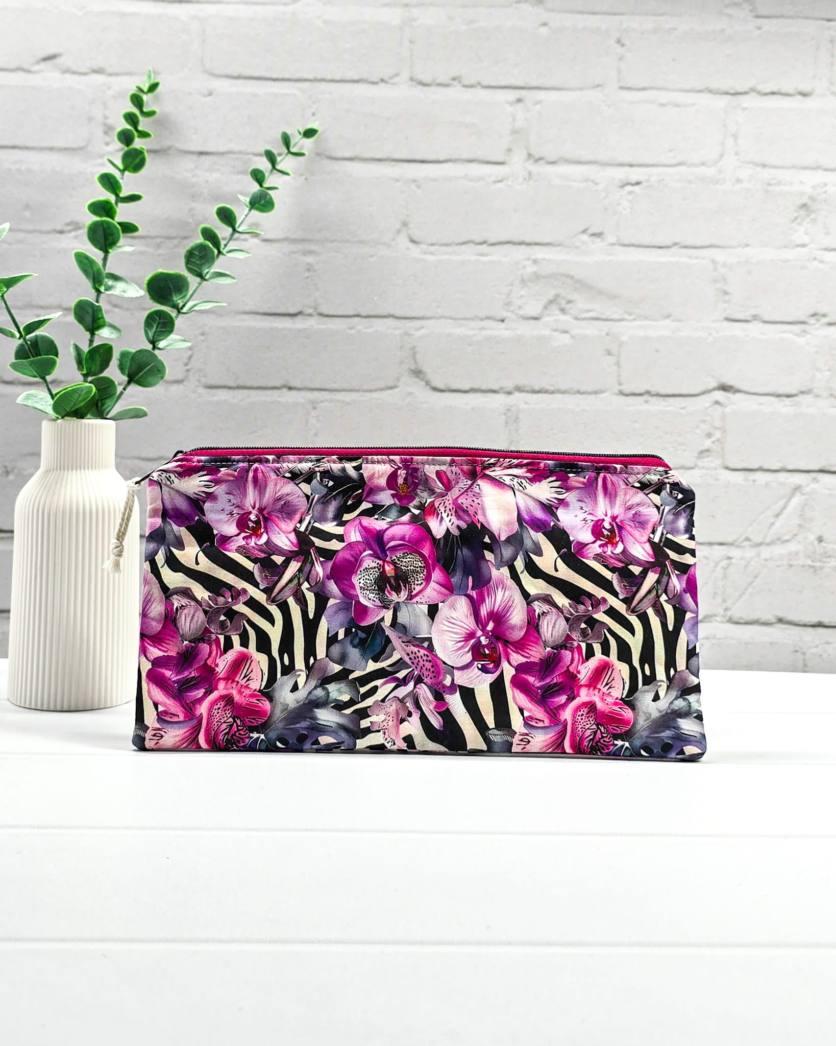 Längliche Tasche mit pinkfarbenem Reißverschluss, hellem Kordel-Zipper und auffälligem Muster aus lila Orchideenblüten über einem schwarz-weißen Zebramuster. Handgenäht in Bayern von Nadelmikado.