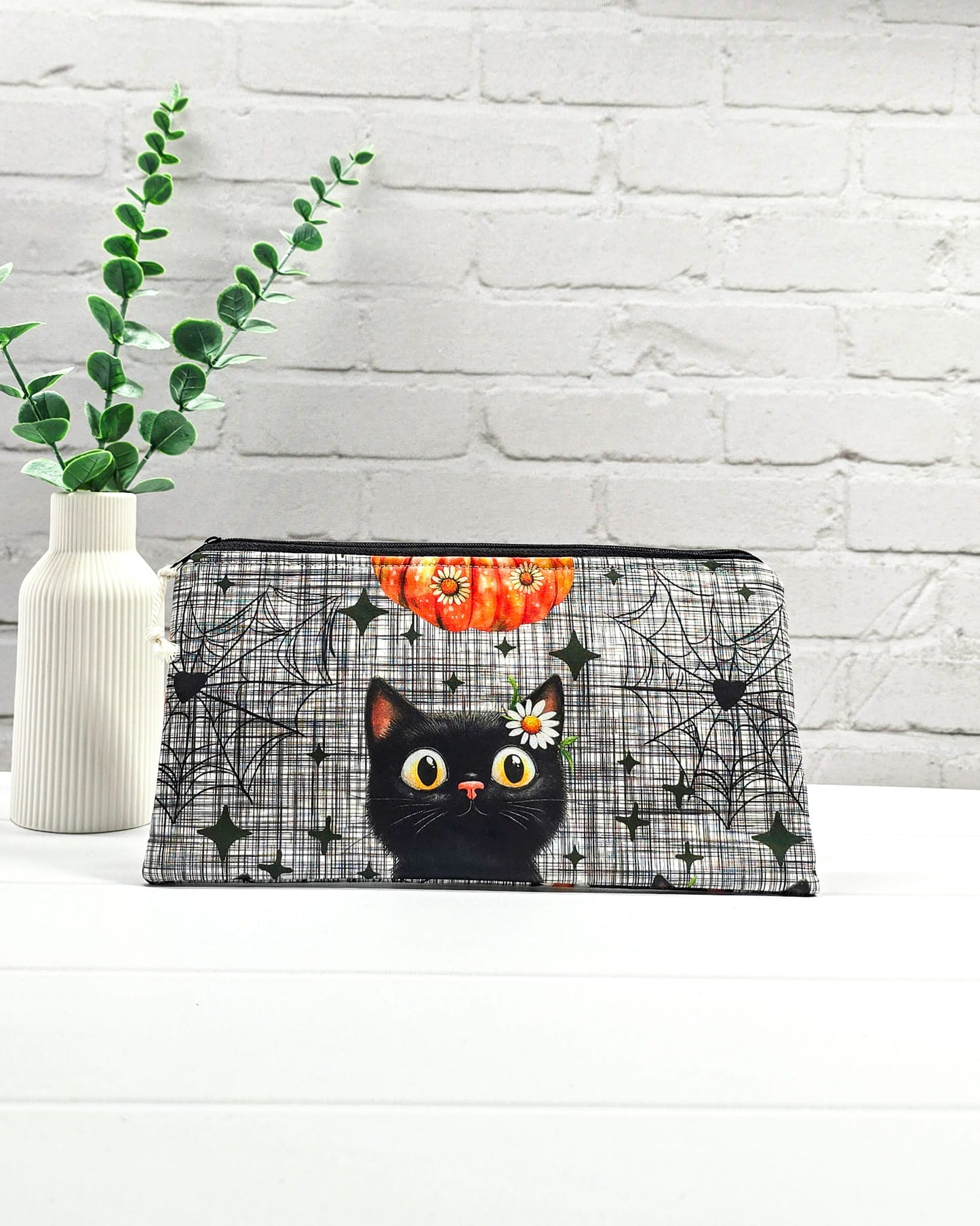 Längliche Zubehörtasche mit schwarzem Reißverschluss, hellem Kordel-Zipper und einer schwarzen Katze vor Spinnweben. Handgenäht in Bayern von Nadelmikado.